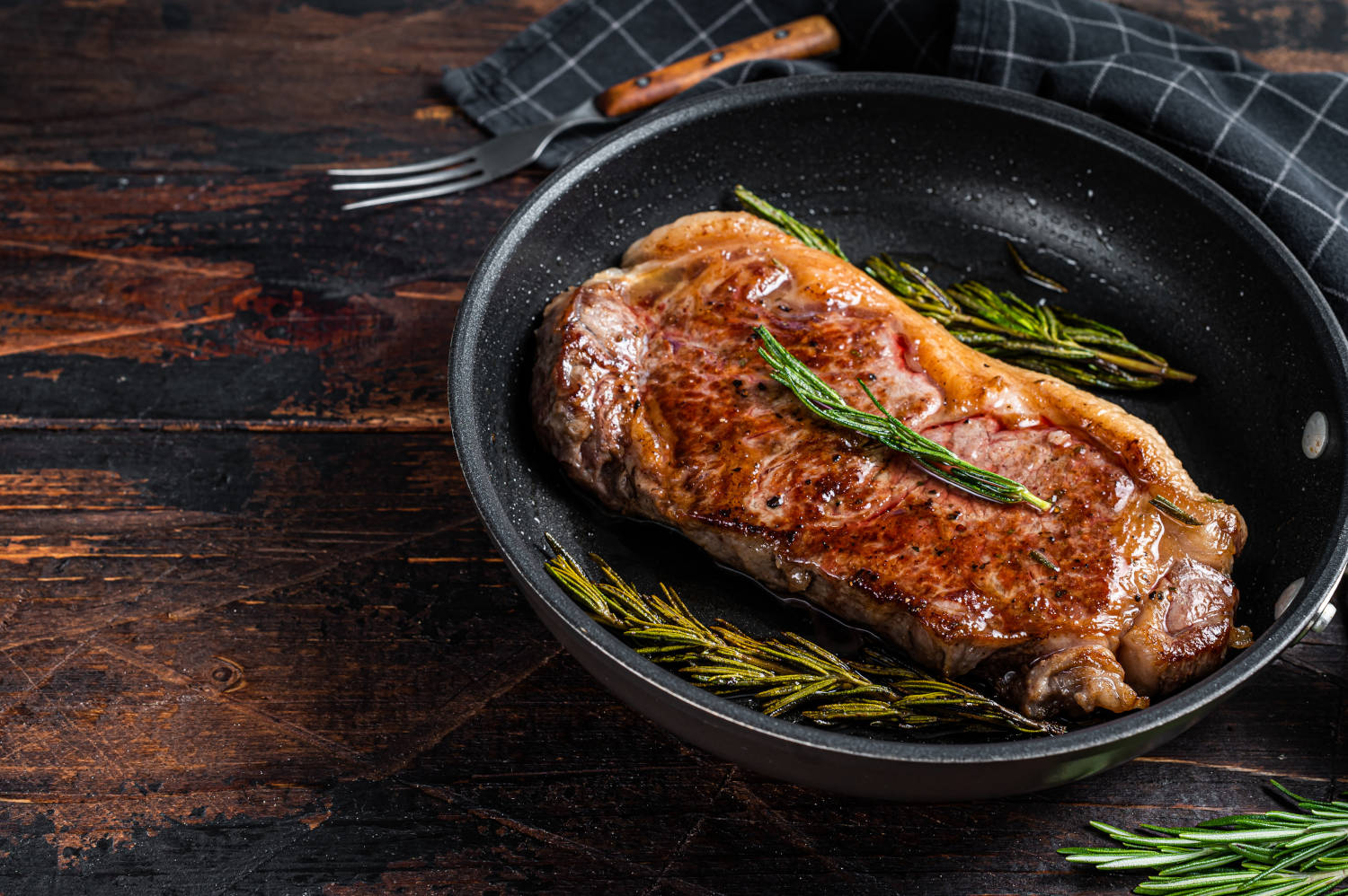 Grilled new york strip beef meat steak in a pan with herbs. Dark wooden background. Top view. Copy space.